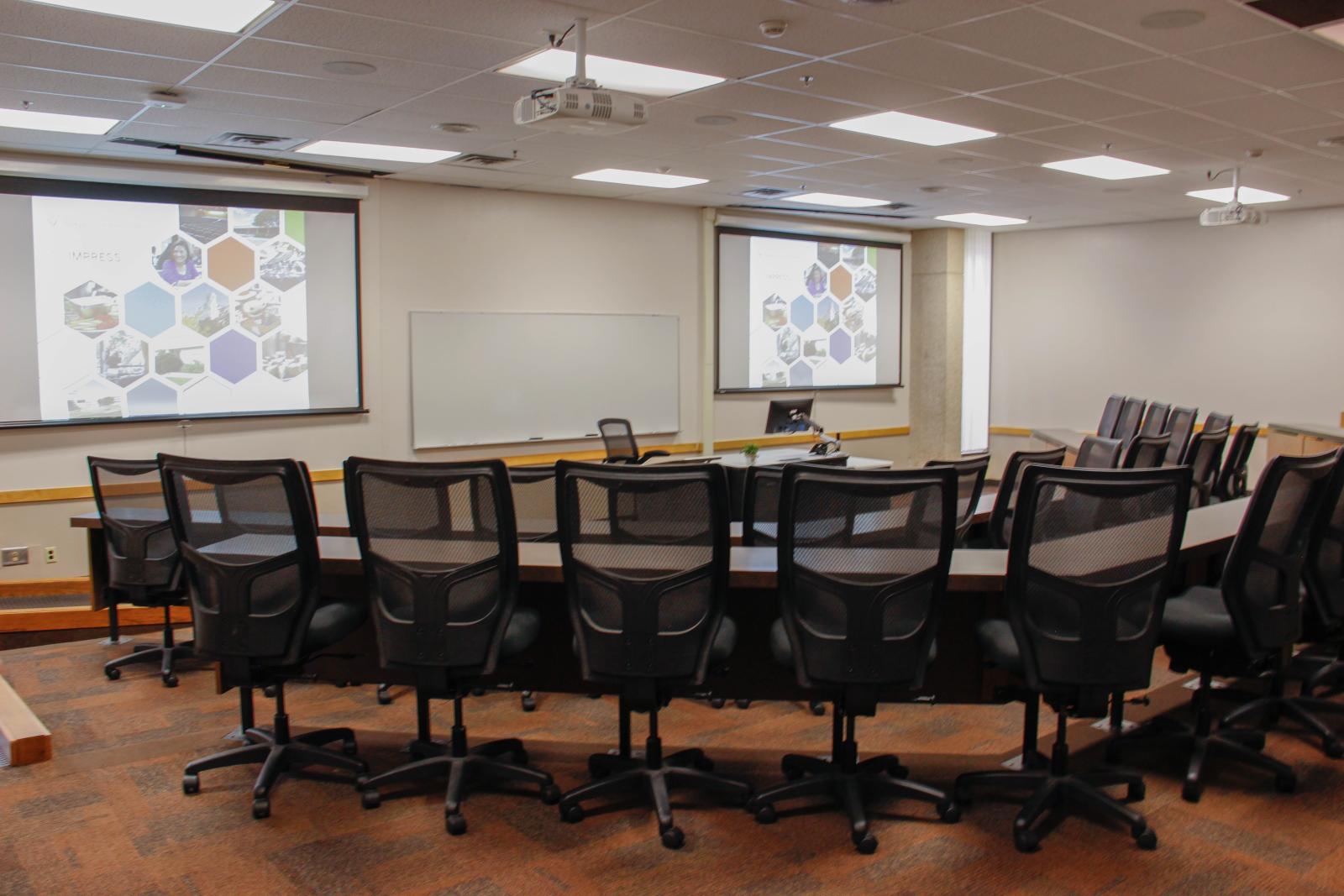 Tiered auditorium-style seating classroom facing a desk, whiteboard and screen.