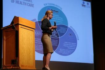 Woman presenting in front of a podium with a large screen displaying a slide with a diagram